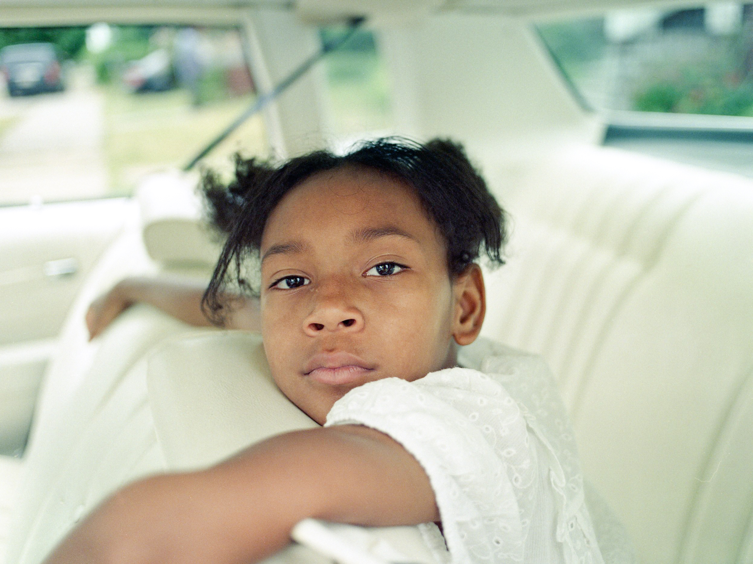 A brightly lit image of a young African American girl in a white dress is sitting in the back seat of a classic car with a white interior. Her arms are draped over the seat in front of her. She is looking directly at the camera with a firm gaze.